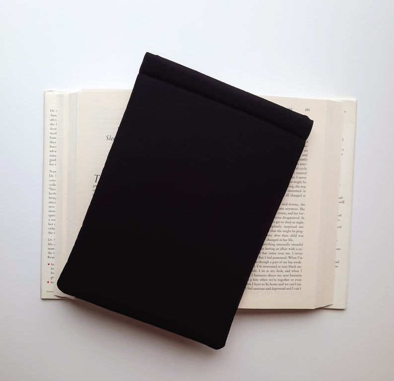 May include: A black book cover rests on an open book with text visible on the pages. The book cover is rectangular and covers a portion of the book. The background is white.