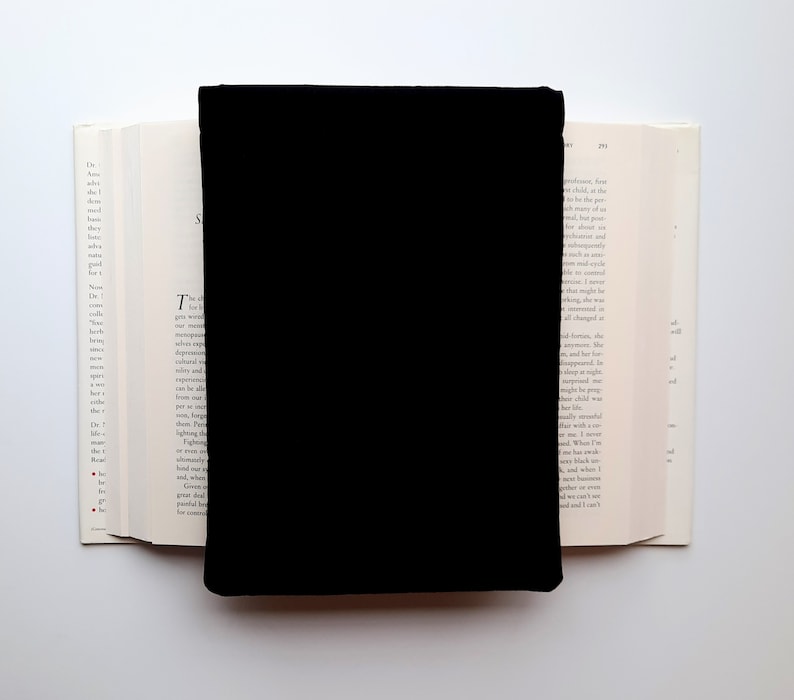 May include: A black book cover is centered on an open book with white pages. The book's pages are visible on either side of the black cover, creating a contrast between the dark cover and the light pages. The background is white.