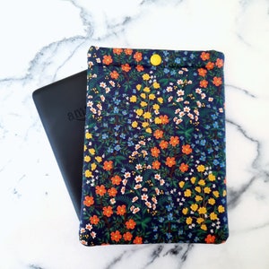 May include: A navy blue e-reader case with a floral pattern of orange, yellow, and white flowers. The case has a yellow button closure. A black e-reader is partially visible next to the case.