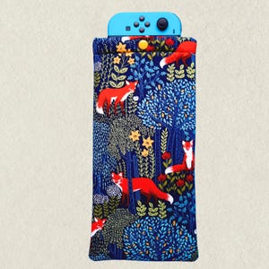 May include: A blue Nintendo Switch case with a forest and fox pattern. The case features a dark blue background with red foxes, yellow flowers, and green foliage. The top of the case holds a blue Switch controller.