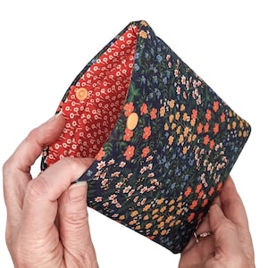 May include: A navy blue floral pouch with a red interior. The exterior features a pattern of small orange, yellow, and white flowers. The pouch is secured with two orange press studs.