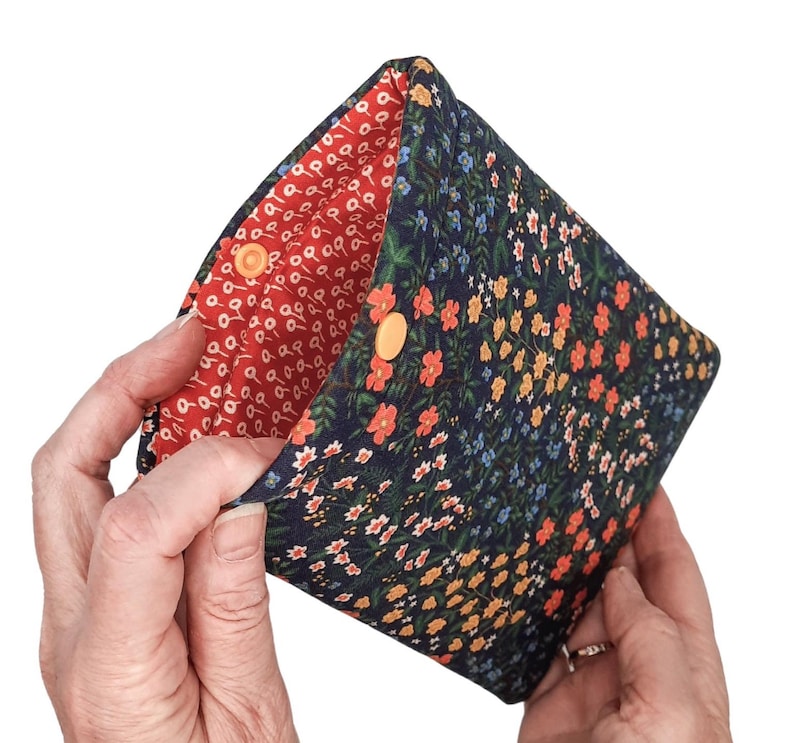 May include: A navy blue floral pouch with a red interior. The exterior features a pattern of small orange, yellow, and white flowers. The pouch is secured with two orange press studs.