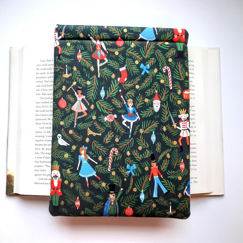 May include: A rectangular fabric book sleeve featuring a dark green background with a repeating pattern of nutcrackers, ballerinas, candy canes, Christmas ornaments, and pine branches. The sleeve is positioned over an open hardcover book.