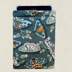 May include: A teal fabric sleeve with a butterfly and floral pattern, designed to hold a Kindle. The sleeve has a button closure at the top. The butterflies are in shades of teal, orange, and white.
