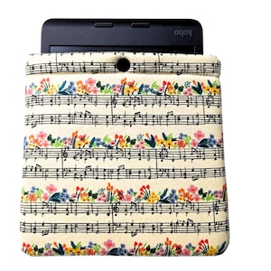 May include: Cream-colored e-reader sleeve with a floral and musical note pattern. The sleeve features a black button closure and is designed to hold an e-reader. The floral design includes pink, blue, and yellow flowers.