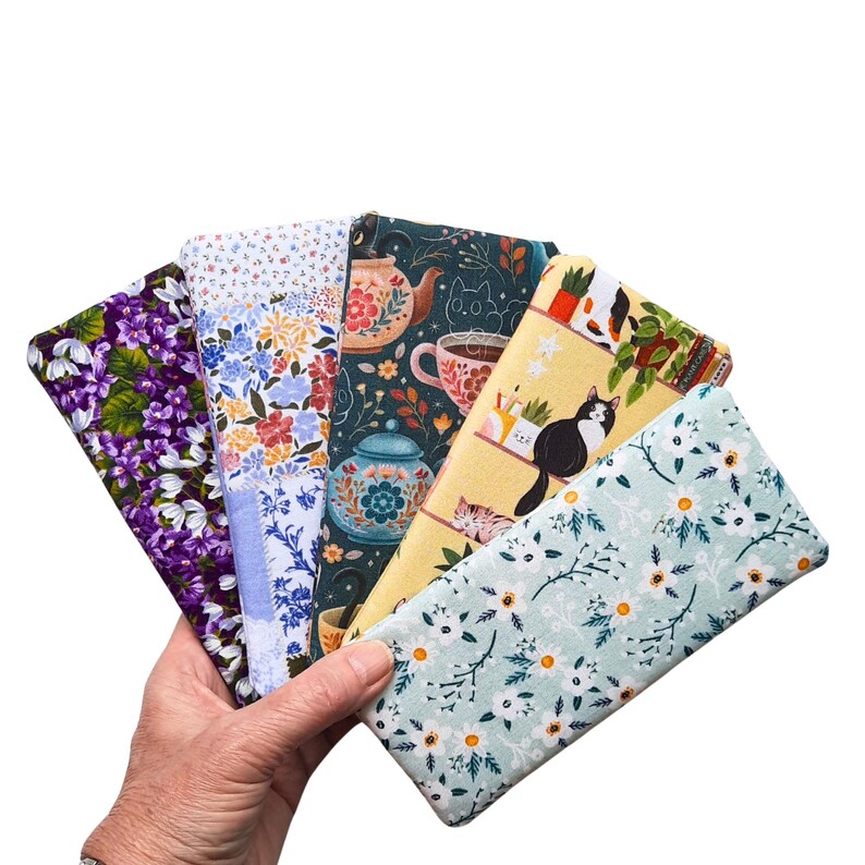 Pu&ograve; includere: Una collezione di pochette in tessuto con vari motivi. I disegni includono fiori, tazze da t&egrave;, teiere e gatti. Le pochette sono tenute in mano, mostrando i diversi motivi e colori. Le pochette sono rettangolari e sembrano essere di cotone.