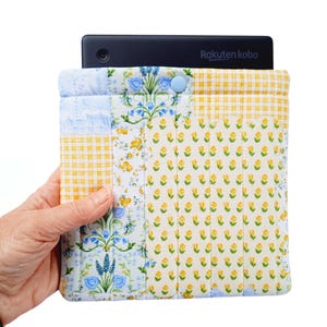May include: A quilted tablet sleeve with a floral and gingham pattern in yellow, blue, and white. The sleeve has a button closure and holds a black Rakuten Kobo device. The design features a patchwork style with various fabric patterns.
