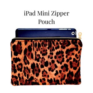 May include: An iPad Mini zipper pouch with a leopard print design. The pouch is tan and brown with a gold zipper and a dark blue iPad Mini inside. The text "iPad Mini Zipper Pouch" is above the pouch.