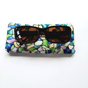 May include: Black sunglasses with brown lenses rest on a colorful, stained-glass patterned case. The case features a vibrant mix of blue, green, purple, and yellow shapes. The sunglasses have a rectangular frame.
