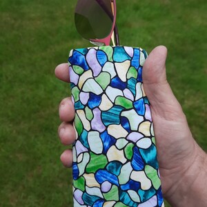 May include: A colorful eyeglasses case with a stained glass pattern in shades of blue, green, and purple. The case holds a pair of sunglasses with a pink frame and green lenses. The case is held in a hand against a green background.