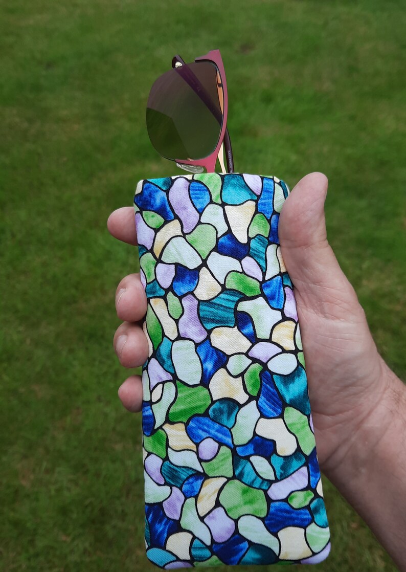 May include: A colorful eyeglasses case with a stained glass pattern in shades of blue, green, and purple. The case holds a pair of sunglasses with a pink frame and green lenses. The case is held in a hand against a green background.