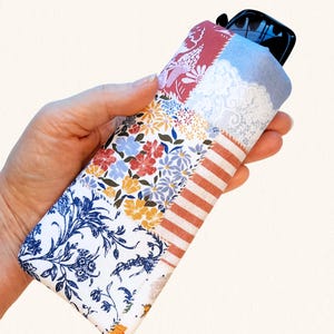 May include: A rectangular fabric pouch featuring a patchwork design with various patterns including blue and white floral motifs, small yellow and red flowers, white lace textures, and orange and white stripes, containing black sunglasses.