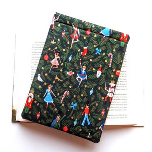 May include: A rectangular fabric sleeve with a black background featuring a repeating pattern of green pine branches, red and blue nutcracker figures, ballerinas, candy canes, stockings, and ornaments. The sleeve is folded over at the top edge.