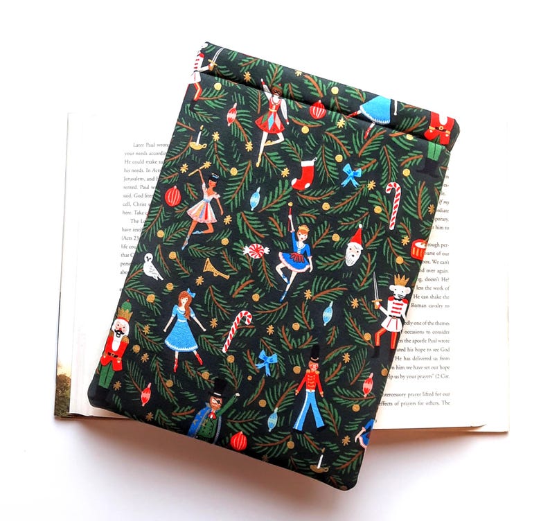 May include: A rectangular fabric sleeve with a black background featuring a repeating pattern of green pine branches, red and blue nutcracker figures, ballerinas, candy canes, stockings, and ornaments. The sleeve is folded over at the top edge.