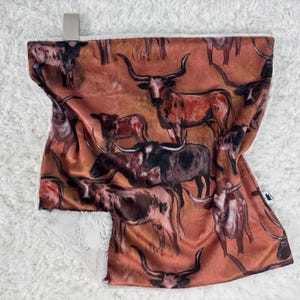 May include: A rectangular blanket featuring a repeating pattern of longhorn cattle in shades of brown, black, and white against a reddish-orange background. The blanket has a soft, white, fluffy backing.