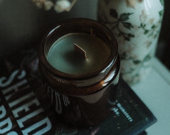 May include: A brown glass jar candle with a wooden wick sits atop a book titled "Shield of Thorns". A small vase with floral patterns and dried flowers is in the background. The candle has a light green wax.