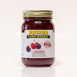 May include: A glass jar of cherry fruit preserves with a gold lid. The label reads "Fowl's Farm Market" and "Cherry Fruit Conserves Net Wt. 20 oz. (567g)". A red sticker with the words "All Natural" is on the label. The jar is sitting on a white surface.
