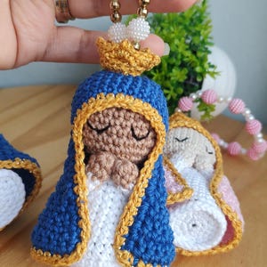 Op de afbeelding: Een gehaakt figuurtje met een bruine huid, gekleed in een blauwe en gouden kapmantel en een gouden kroon, met de handen gevouwen in gebed. Het figuurtje is bevestigd aan een snoer van witte en gouden kralen. Een tweede figuurtje is zichtbaar.