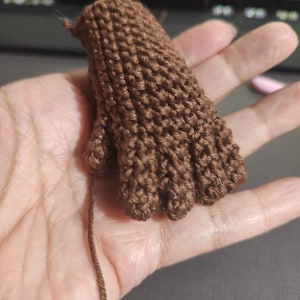 May include: A brown knitted hand with five fingers. The hand is made of yarn and is being held by a person's hand.