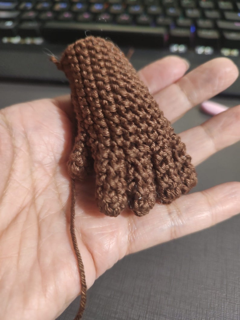 May include: A brown knitted hand with five fingers. The hand is made of yarn and is being held by a person's hand.