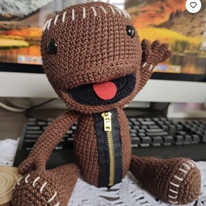 May include: A handmade brown crochet doll with a black mouth, red tongue, and a gold zipper. The doll has black button eyes and white stitching details. The doll is waving its right hand.