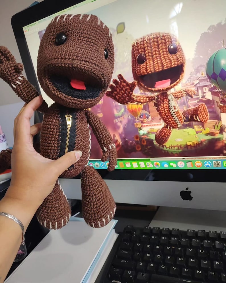 May include: A brown crocheted LittleBigPlanet Sackboy toy with a zipper and a red tongue. The toy is standing on a table in front of a computer screen showing a video game character.