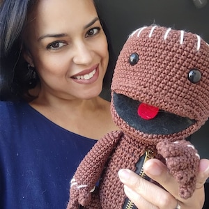 May include: A woman is holding a brown crocheted plush toy with a zipper and a red tongue. The toy has black eyes and a stitched smile.