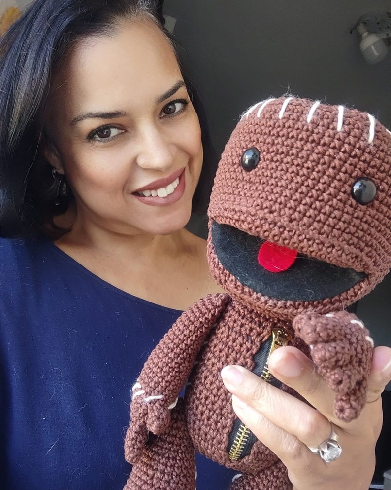May include: A woman is holding a brown crocheted plush toy with a zipper and a red tongue. The toy has black eyes and a stitched smile.
