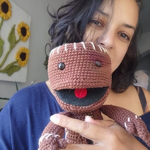 May include: A brown crocheted plush toy with a stitched smile and red tongue. The toy has black eyes and a stitched scar on its face. The toy is being held by a person.