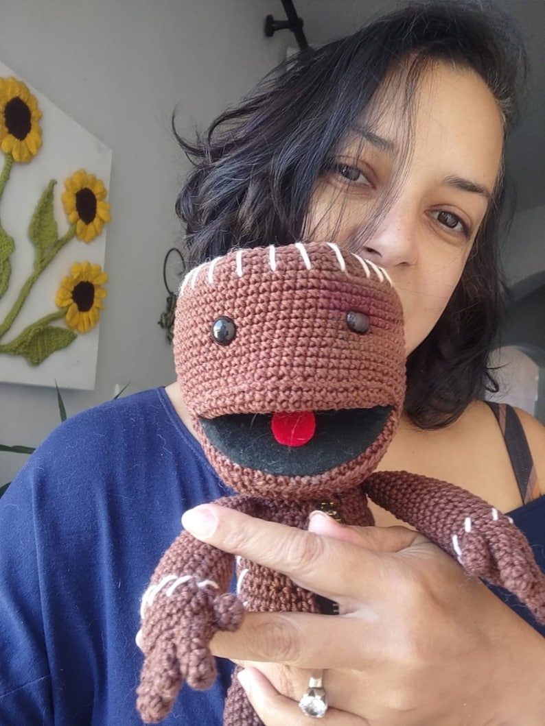 May include: A brown crocheted plush toy with a stitched smile and red tongue. The toy has black eyes and a stitched scar on its face. The toy is being held by a person.