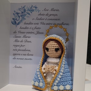 Op de afbeelding: Een gehaakt beeldje van de Maagd Maria in een blauwe en witte jurk met een gouden rand. Ze houdt een witte rozenkrans vast met parel kralen. Het beeldje is ingelijst in een wit frame met een geprint gebed in het Portugees.