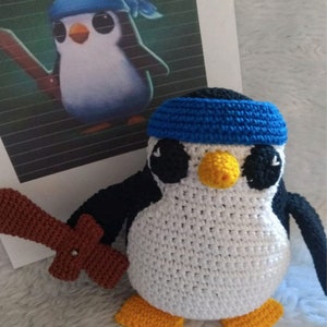 Puede incluir: Un juguete de pingüino de crochet que lleva una diadema azul y sostiene una espada de madera marrón. El pingüino tiene ojos negros, un pico amarillo y plumas blancas y negras.