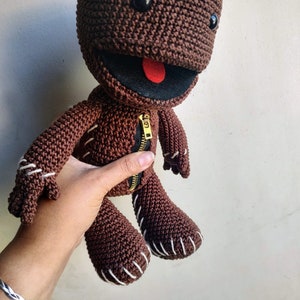 May include: Brown crocheted plush toy with a zipper down the front and a red tongue sticking out. The toy has black eyes and white stitching around the edges.