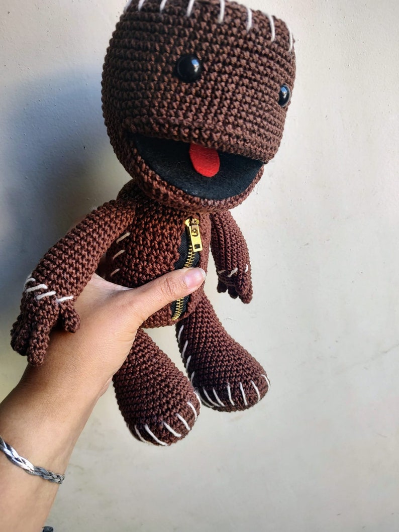 May include: Brown crocheted plush toy with a zipper down the front and a red tongue sticking out. The toy has black eyes and white stitching around the edges.