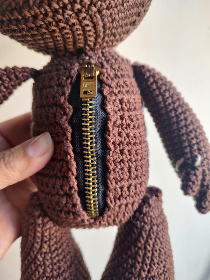 May include: A brown crocheted plush toy with a gold zipper down the back. The zipper has the word "OPTI" printed on it.