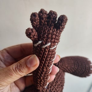 May include: A brown crocheted hand with five fingers, stitched with white thread. The hand is attached to a brown crocheted body.