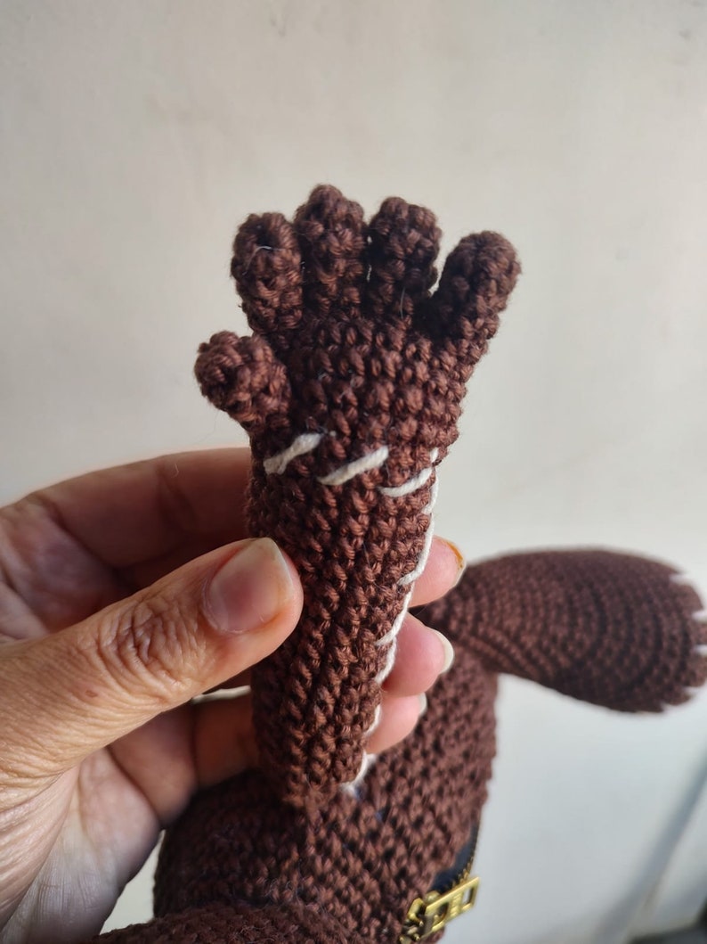 May include: A brown crocheted hand with five fingers, stitched with white thread. The hand is attached to a brown crocheted body.