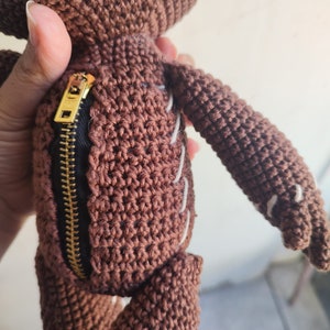 May include: A brown crocheted plush toy with a zipper on the side. The toy has white stitching details and a gold zipper pull.