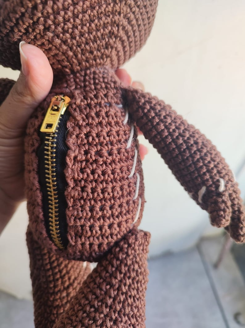 May include: A brown crocheted plush toy with a zipper on the side. The toy has white stitching details and a gold zipper pull.