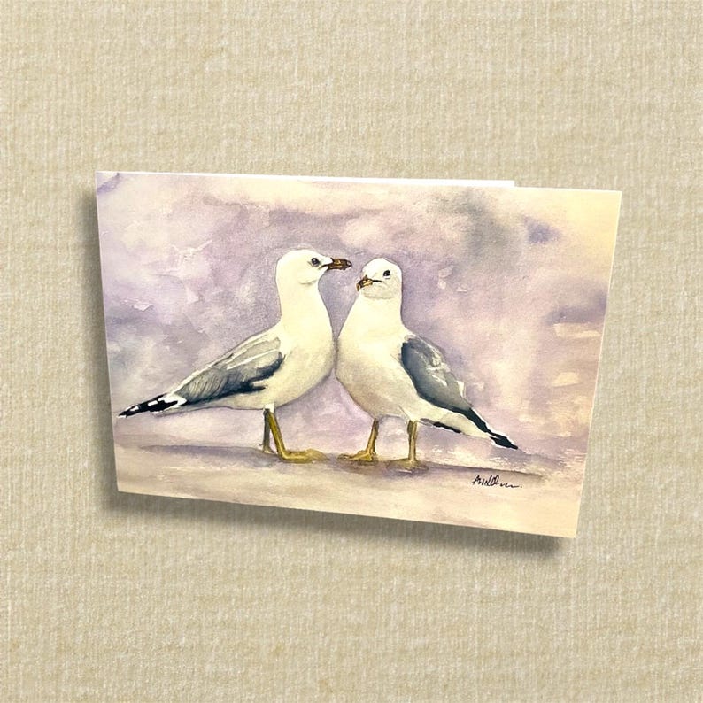May include: Watercolor painting of two seagulls standing close together. The birds have white bodies, gray wings, and yellow legs. The background is a soft blend of purple and gray, creating a serene coastal scene.