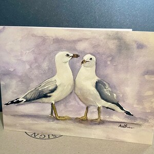 Watercolour print painting of two white seagulls with grey wings, facing each other on a purple background. The artist's signature, is in lower right corner.