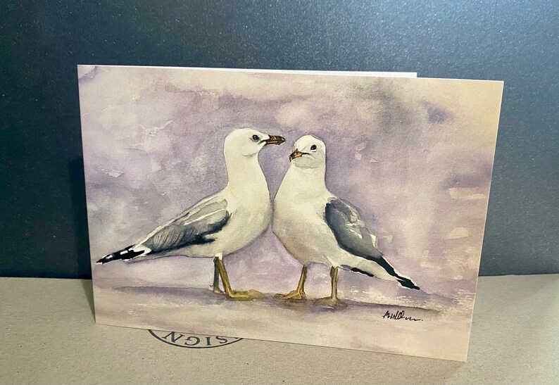 Watercolour print painting of two white seagulls with grey wings, facing each other on a purple background. The artist's signature, is in lower right corner.