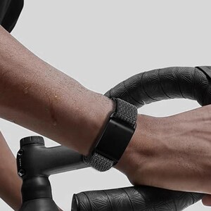May include: A black digital fitness tracker with a textured grey fabric band worn on the wrist. The device features a rectangular screen and is positioned while gripping black bicycle handlebars with a patterned grip tape finish.