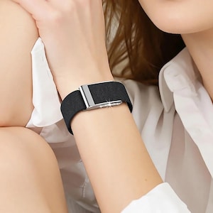 May include: A black elastic watch band featuring a rectangular silver-toned metal buckle. The buckle is accented with a row of small, clear, sparkling stones along the side, providing a decorative detail to the simple, solid-colored fabric strap.