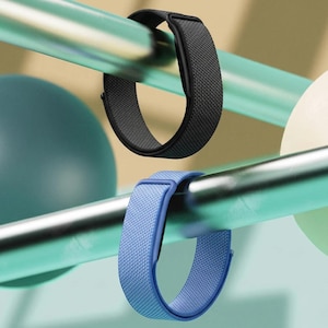 May include: Two watch bands, one black and one light blue, are displayed hanging from horizontal bars. Each band features a woven fabric texture and a matching plastic connector piece, showcasing a simple, functional design for a wearable device.