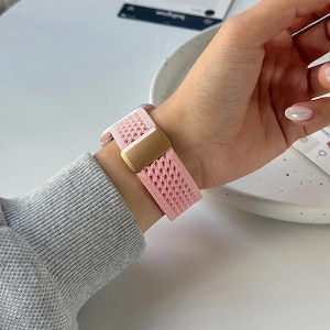 May include: A pink watch band with a woven design and a gold-colored clasp is fastened on a wrist. The band is made of a soft, breathable material. The watch band is a stylish accessory.