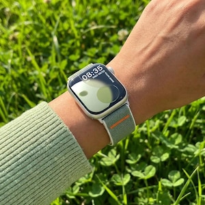May include: A silver smartwatch with a green and orange striped band displays the time 08:35. The watch face shows a green and white design. The watch is worn on a wrist, with a green sweater sleeve visible in the foreground.