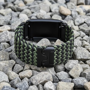 May include: A watch band featuring a woven paracord design in alternating black and olive green colors. The band includes a matte black metal buckle mechanism and black metal connectors for attaching to a watch face, creating a durable look.