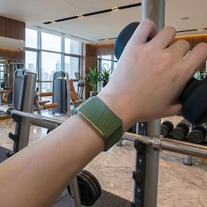 May include: A smartwatch with a matte olive green silicone band and a rectangular metallic frame, worn on the wrist while gripping a black gym weight. The watch features a smooth, solid-colored strap and a clean, minimalist design profile.