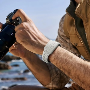 May include: A light gray silicone watch band featuring a textured, rugged link design and a rectangular face cover with a prominent embossed letter L logo, worn on a wrist while holding a black professional camera with a Zeiss lens.